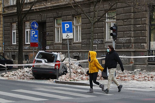 A couple walk past a car damaged by the earthquake in downtown Zagreb, Croatia, Monday, March 23, 2020. Croatia experienced a series of aftershocks a day after a strong earthquake triggered widespread damage and injured over 20 people in the capital. (AP Photo/Darko Bandic)