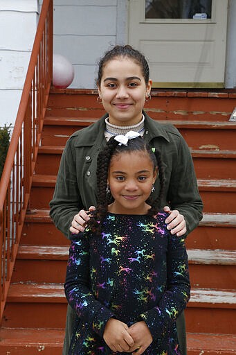 Jalen Grimes, 13, stands behind 7-year-old sister, Sydney, outside their home in Chicago on Sunday, March 22, 2020. Both girls are now doing schoolwork at home because of the virus pandemic. Jalen is missing much of her 8th grade year, including a class trip. Usually 8th graders in Illinois have a graduation, a rite of passage into high school. But those ceremonies, which often happen in June, are a question mark, as well. Jalen says she is annoyed, sad and worried all at once. (AP Photo/Martha Irvine)