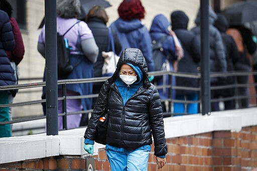 A medical worker wearing a single protective glove and a face mask walks past a line of workers and visitors waiting to be tested for COVID-19, the disease caused by the new coronavirus, at the main entrance to the Department of Veterans Affairs Medical Center, Monday, March 23, 2020, in New York.  (AP Photo/John Minchillo)