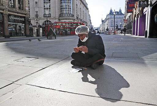 An employee of a nearby takeaway restaurant rolls a cigarette as he has a break sitting on the pavement near Piccadilliy Circus in central London, Tuesday, March 24, 2020.  Britain's Prime Minister Boris Johnson on Monday imposed its most draconian peacetime restrictions due to the spread of the coronavirus on businesses and social gatherings. For most people, the new coronavirus causes only mild or moderate symptoms. For some it can cause more severe illness.(AP Photo/Frank Augstein)