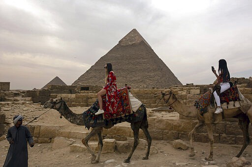 In this Tuesday, March 10, 2020, photo, two foreign women tourists ride camels at the Giza Pyramids near Cairo, Egypt. The country's prime minister announced at a news conference on Tuesday, March 24, 2020, a two-week, 7 p.m. to 6 a.m. curfew for its over 100 million people to slow the spread of the new coronavirus. The 11-hour curfew would go into effect Wednesday across the country and last for two weeks. (AP Photo/Maya Alleruzzo)