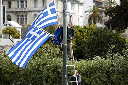 Municipal workers install Greek flags in front of the Greek parliament in Athens on Tuesday, March 24, 2020. An annual military parade has been canceled Wednesday amid a circulation ban imposed to slow the spread of the coronavirus but a flyover with jets and helicopters will take place. For some people the new COVID-19 coronavirus causes only mild or moderate symptoms, but for some it can cause severe illness including pneumonia. (AP Photo/Thanassis Stavrakis)