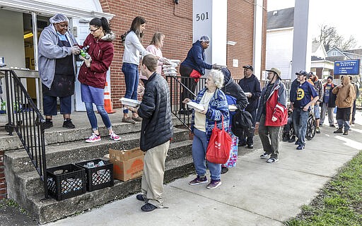 A line forms at the front steps of the Daniel Pitino Shelter as shelter staff serve carry out lunch meals to visitors, Wednesday, March 25, 2020, in Owensboro, Ky. Lunch is normally served in the shelter's dining area but carry out is now the current protocol for the shelter during the coronavirus pandemic. (Greg Eans/The Messenger-Inquirer via AP)