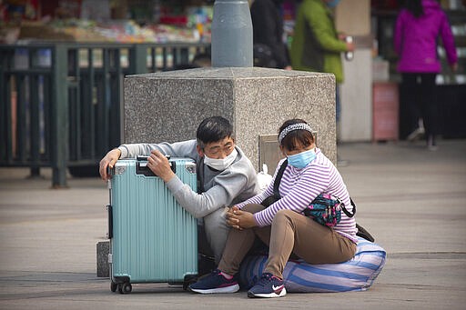 Travelers wearing face masks sit with their luggage outside the Beijing Railway Station in Beijing, Wednesday, March 25, 2020. Some train stations and bus services reopened in China's Hubei Province on Wednesday and people who passed a health check would finally be allowed to travel for the first time since the coronavirus outbreak surged in January. The new coronavirus causes mild or moderate symptoms for most people, but for some, especially older adults and people with existing health problems, it can cause more severe illness or death. (AP Photo/Mark Schiefelbein)