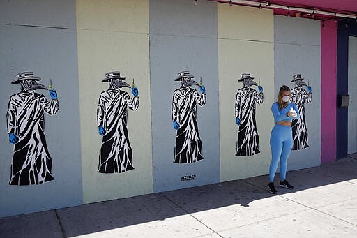 Arianna Gnagnarelli puts on gloves and wears a mask before having friends take a picture of her at a closed and boarded up business due to coronavirus Wednesday, March 25, 2020, in Las Vegas. Local artists have been painting the raw plywood boards on shuttered businesses in the area know as the Arts District. (AP Photo/John Locher)