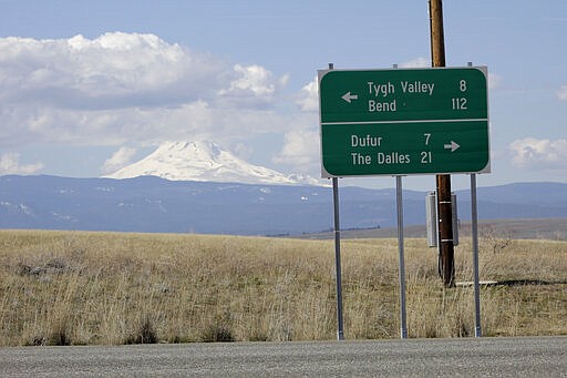 In this photo taken March 20, 2020, a sign indicates the distance to the nearest towns as cloud-capped Mount Hood rises in the background in this photo taken near Dufur, Oregon. Tiny towns tucked into Oregon's windswept plains and cattle ranches miles from anywhere in South Dakota might not have had a single case of the new coronavirus yet, but their residents fear the spread of the disease to areas with scarce medical resources, the social isolation that comes when the only diner in town closes its doors and the economic free fall that's already hitting them hard. (AP Photo/Gillian Flaccus)