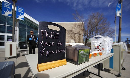 A sign encourages young people to pick up a lunch outside the Aurora Public Library as branch manager Phillip Challis looks on in an effort to help residents and reduce the spread of the new coronavirus Wednesday, March 25, 2020, in Aurora, Colo. The new coronavirus causes mild or moderate symptoms for most people, but for some, especially older adults and people with existing health problems, it can cause more severe illness or death. (AP Photo/David Zalubowski)