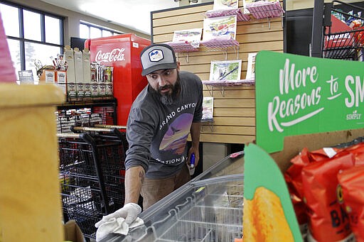 In this photo taken March 20, 2020, Mike Johnston, a clerk at the Maupin Market in tiny Maupin, Oregon, wipes down the ice cream case to protect customers from the new coronavirus. Tiny towns tucked into Oregon's windswept plains and cattle ranches miles from anywhere in South Dakota might not have had a single case of the new coronavirus yet, but their residents fear the spread of the disease to areas with scarce medical resources, the social isolation that comes when the only diner in town closes its doors and the economic free fall that's already hitting them hard. (AP Photo/Gillian Flaccus)