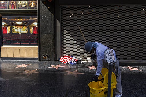 A worker performs a daily cleaning of the sidewalk of the closed Hollywood Pantages Theatre on the Hollywood Blvd., as a homeless person sleeps under a blanket in Los Angeles Tuesday, March 24, 2020. (AP Photo/Damian Dovarganes)