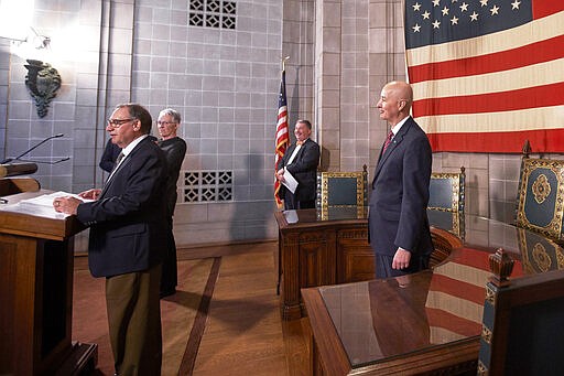 In this March 20, 2020 photo, participants in a press briefing on the Covid-19 virus in Lincoln, Neb., from left: Neb. Chief Medical Officer Dr. Gary Anthone, the sign language interpreter, Jim Macy, Director of the Nebraska Department of Environment and Energy and Gov. Pete Ricketts, maintain social distancing. Dr. Anthone is holding on to the lectern as will all the other participants in the news conference. (AP Photo/Nati Harnik)