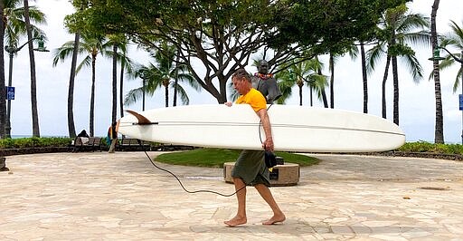 A surfer carries his board along Waikiki Beach in Honolulu on Thursday, March 26, 2020. A 14-day quarantine on travelers takes effect in Hawaii, one of the world's most famous tourist destinations, as the island state fights the coronavirus. Hawaii welcomed 10 million travelers last year, for an average of about 830,000 a month. Authorities expect this number will dwindle to nearly zero in April as the new rule is implemented. Dozens of hotels are temporarily shutting their doors in response. (AP Photo/Caleb Jones)