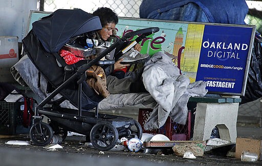 In this Tuesday, March 17, 2020 photo, a homeless person holds a shoe while camped on a street bench in Oakland, Calif. Efforts to move California's large homeless population into isolated quarters to slow the spread of the coronavirus are still sluggish despite a $150 million cash infusion by the governor and a crisis that public health experts say needs to be curbed now.  (AP Photo/Ben Margot)