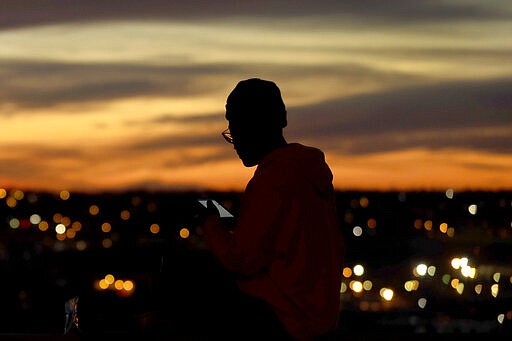 A person looks at a smart device as the sun sets, Wednesday, March 25, 2020, in Kansas City, Mo. The city along with neighboring counties is under Stay at Home orders to help prevent the spread of COVID-19, the disease caused by the new coronavirus. (AP Photo/Charlie Riedel)