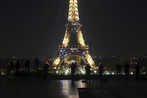 Media stand in front of the Eiffel Tower where the word &quot;Merci&quot;, the French word for 'Thank you&quot;, is emblazoned as France's coronavirus death toll continued to climb, in Paris, Friday, March 27, 2020. Health workers fighting to save lives in France from COVID-19 have received a huge show of gratitude, from the Eiffel Tower. The new coronavirus causes mild or moderate symptoms for most people, but for some, especially older adults and people with existing health problems, it can cause more severe illness or death. (AP Photo/Thibault Camus)