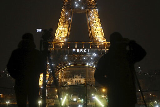 Cameramen stand in front of the Eiffel Tower where the word &quot;Merci&quot;, the French word for 'Thank you&quot;, is emblazoned as France's coronavirus death toll continued to climb, in Paris, Friday, March 27, 2020. Health workers fighting to save lives in France from COVID-19 have received a huge show of gratitude, from the Eiffel Tower. The new coronavirus causes mild or moderate symptoms for most people, but for some, especially older adults and people with existing health problems, it can cause more severe illness or death. (AP Photo/Thibault Camus)