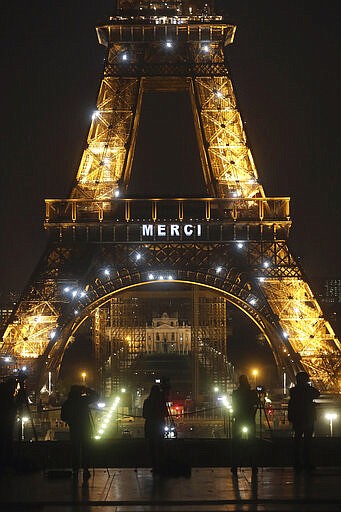 Media stand in front of the Eiffel Tower where the word &quot;Merci&quot;, the French word for 'Thank you&quot;, is emblazoned as France's coronavirus death toll continued to climb, in Paris, Friday, March 27, 2020. Health workers fighting to save lives in France from COVID-19 have received a huge show of gratitude, from the Eiffel Tower. The new coronavirus causes mild or moderate symptoms for most people, but for some, especially older adults and people with existing health problems, it can cause more severe illness or death. (AP Photo/Thibault Camus)