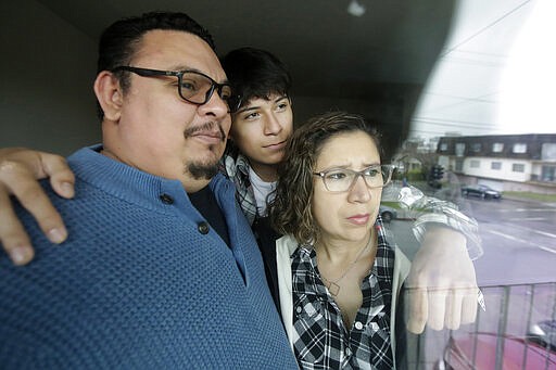 In this Tuesday, March 24, 2020, photo, William Gonzalez, from left, poses for photos with son Ricardo Bautista, 14, and wife Sonia Bautista through a window of their apartment in South San Francisco, Calif. California's Bay Area has been shut down for more than a week, the first region of America to order its residents to stay home, work remotely and homeschool their children in a desperate bid to slow the spread of the coronavirus pandemic. (AP Photo/Jeff Chiu)