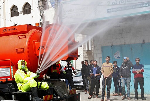 In this March 27, 2020, photo, residents watch workers wearing protective gear spray disinfectant as a precaution against the coronavirus, at the main road of closed market of Shijaiyah neighborhood in Gaza City. Gaza municipality closed all the weekly Friday markets in Gaza starting from today. (AP Photo/Adel Hana)