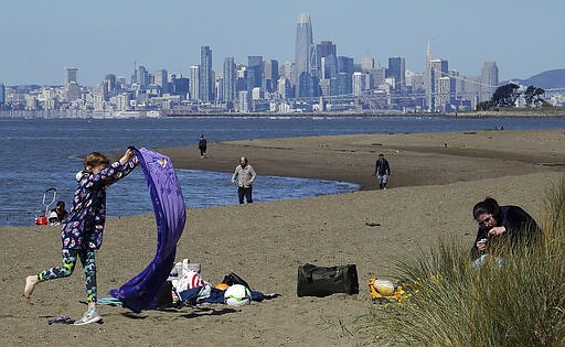 FILE - In this March 26, 2020, file photo, a girl lays out a blanket at Robert W. Crown Memorial State Beach in Alameda, Calif. All over the country, outdoor spots have only grown more crowded since stores, restaurants and schools have closed their doors and officials have issued stay-at-home orders. In the background is the skyline of San Francisco. (AP Photo/Ben Margot File)