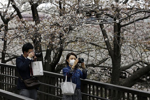 Masked people stop on a bridge to photograph cherry blossoms Friday, March 27, 2020, in Tokyo. The new coronavirus causes mild or moderate symptoms for most people, but for some, especially older adults and people with existing health problems, it can cause more severe illness or death. (AP Photo/Kiichiro Sato)