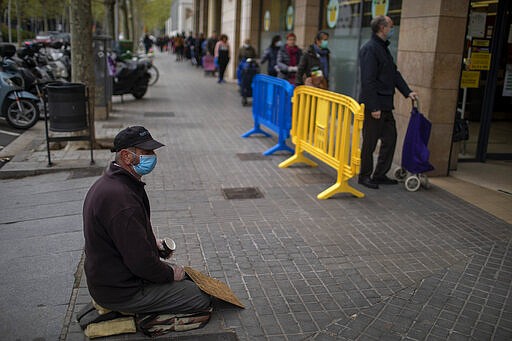 A man begs for alms as people line up to buy supplies from a shop during the coronavirus outbreak in Barcelona, Spain, Friday, March 27, 2020. The new coronavirus causes mild or moderate symptoms for most people, but for some, especially older adults and people with existing health problems, it can cause more severe illness or death. (AP Photo/Emilio Morenatti)