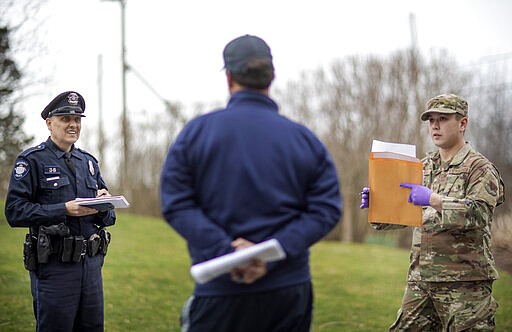 Rhode Island Air National Guard TSgt. William Randall, right, and Westerly police officer Howard Mills talk with New York resident Reha Kocatas who is self-quarantining at his home in Westerly, R.I., Saturday, March 28, 2020. The Rhode Island National Guard started going door to door on Saturday in coastal areas to inform any New Yorkers who may have come to the state that they must self-quarantine for 14 days. (AP Photo/David Goldman)