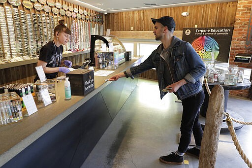 In this photo taken March 24, 2020, Christopher Ellis, right, leans in to show his identification to budtender Maegan Clark, left, as he stands on a line taped on the floor to designate a social distancing separation due to the new coronavirus, while shopping at The Reef Capitol Hill, a marijuana store in Seattle. Earlier in the week, Washington Gov. Jay Inslee ordered nonessential businesses to close and the state's more than 7 million residents to stay home in order to slow the spread of the new coronavirus. In Washington and several other states where marijuana is legal, pot shops and workers in the market's supply chain were deemed essential and allowed to remain open. (AP Photo/Ted S. Warren)