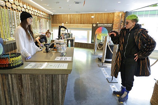 In this photo taken March 24, 2020, Ray Ray, right, talks with budtender Peyton Beech, left, as he stands on a line taped on the floor to designate a social distancing separation due to the new coronavirus while shopping at The Reef Capitol Hill, a marijuana store in Seattle. Earlier in the week, Washington Gov. Jay Inslee ordered nonessential businesses to close and the state's more than 7 million residents to stay home in order to slow the spread of the new coronavirus. In Washington and several other states where marijuana is legal, pot shops and workers in the market's supply chain were deemed essential and allowed to remain open. (AP Photo/Ted S. Warren)