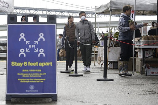 A sign is shown advising people to maintain distance is shown as a man wears a mask while waiting in line at the Ferry Plaza Farmers Market in San Francisco, Saturday, March 28, 2020. (AP Photo/Jeff Chiu)