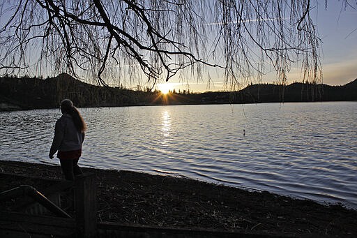 In this March 3, 2020, photo, Barbara Austin, a homeowner who lives along the edge of the reservoir created by the Copco 1 Dam, walks along the shore at sunset near Copco, Calif. A plan to demolish four dams on California's second-largest river to benefit threatened salmon has sharpened a decades-old dispute over who has the biggest claim to the river's life-giving waters. The project, if it goes forward, would be the largest dam demolition project in U.S. history and would drain the reservoir. Residents who live on its shores are fighting the plan. (AP Photo/Gillian Flaccus)