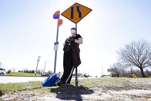 Maryam Jorgensen, of Des Moines, Iowa, wears gloves and a face mask as she waits to catch a bus after grocery shopping, Monday, March 30, 2020, in Des Moines, Iowa. The new coronavirus causes mild or moderate symptoms for most people, but for some, especially older adults and people with existing health problems, it can cause more severe illness or death. (AP Photo/Charlie Neibergall)