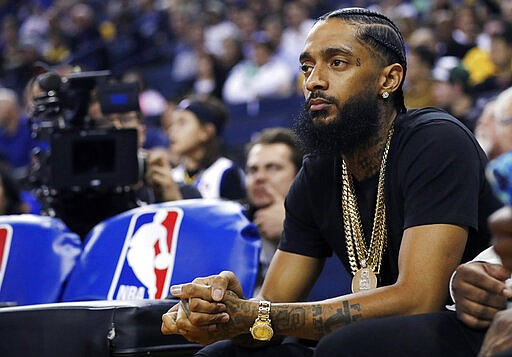 FILE - In this March 29, 2018, file photo, rapper Nipsey Hussle watches an NBA basketball game between the Golden State Warriors and the Milwaukee Bucks in Oakland, Calif. Hussle, 33, was shot and killed outside his Los Angeles clothing store on March 31, 2019. A year after Hussle's death, his popularity and influence are as strong as ever. He won two posthumous Grammys in January, he remains a favorite of his hip-hop peers and his death has reshaped his hometown of Los Angeles in some unexpected ways. (AP Photo/Marcio Jose Sanchez, File)