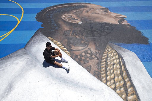 FILE - In this April 17, 2019 file photo, mural artist Gustavo Zermeno Jr. poses for photos on a basketball court mural he dedicated to slain rapper Nipsey Hussle in Los Angeles.  Hussle, 33, was shot and killed outside his Los Angeles clothing store on March 31, 2019. A year after Hussle's death, his popularity and influence are as strong as ever. He won two posthumous Grammys in January, he remains a favorite of his hip-hop peers and his death has reshaped his hometown of Los Angeles in some unexpected ways. (AP Photo/Jae C. Hong, File)