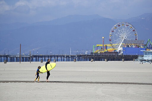 A surfer with a child walks on the mostly empty Santa Monica beach Sunday, March 29, 2020, in Los Angeles. With cases of coronavirus surging and the death toll increasing, lawmakers are pleading with cooped-up Californians to spend a second weekend at home to slow the spread of the infections. (AP Photo/Marcio Jose Sanchez)