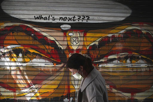 A woman wearing a face mask to protect against coronavirus, passes in front of graffiti painted on the shutter of a closed restaurant during a lockdown order by the Greek government to control the spread of the virus, in Athens, Monday, March 30, 2020. Greece's prime minister is calling on all his cabinet ministers and the lawmakers of his center-right New Democracy party to donate 50% of their salaries over the next two months to the fight against the spread of the new coronavirus. (AP Photo/Thanassis Stavrakis)