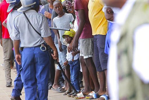 Zimbabwean police stand with people in a queue obeying the rules to prevent the spread of the coronavirus, to enter a supermarket in Harare, Zimbabwe, Monday, March, 30, 2020. Zimbabwe went into a lockdown for 21 days in an effort to curb the spread of the coronoavirus. The new coronavirus causes mild or moderate symptons for most people, but for some, especially older adults and people with existing health problems,it can cause more severe illness or death. (AP Photo/Tsvangirayi Mukwazhi)