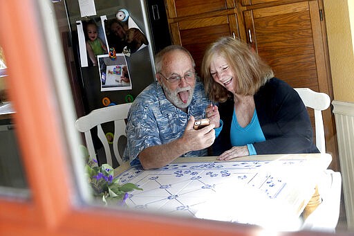 Seen through their kitchen window, Allan and Debbie Cameron contact their grandchildren via the internet Wednesday, March 25, 2020, in Chandler, Ariz. Debbie, 68, has asthma which makes her one of the people most at risk from the new coronavirus. The Cameron's now she see their children and grandchildren from the other side of a window or a phone. (AP Photo/Matt York)
