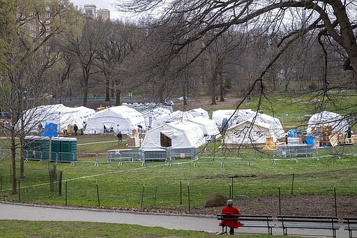 A woman watches from a park bench as the Samaritan's Purse crew and medical personnel work on preparing to open a 68 bed emergency field hospital specially equipped with a respiratory unit in New York's Central Park, Tuesday, March 31, 2020, in New York. The new coronavirus causes mild or moderate symptoms for most people, but for some, especially older adults and people with existing health problems, it can cause more severe illness or death. (AP Photo/Mary Altaffer)