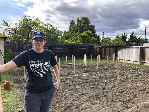 In this March 23, 2020 photo, Lindsay Waldrop stands near her garden at her home in Anaheim, Calif. Waldrop plowed 1,000 square feet of grass to start a garden this year. She has planted dozens of tomatoes, eggplants and peppers with many more crops started as seeds. As the arrival of spring in the Northern Hemisphere coincides with orders to stay at home and out of crowds, the backyard garden has become a getaway for the mind in chaotic times. (Matt Snyder via AP).