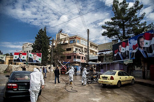 FILE - In this March 24, 2020 file photo, workers spray disinfectant to prevent the spread of the coronavirus, on a street lined with billboards showing Syrian President Bashar Assad, in Qamishli, Syria. In some cells in Iran, Syria and other countries in the Middle East, prisoners are crammed in by the dozens, with little access to hygiene or medical care. So if one infection gets in, the novel coronavirus could run rampant. Among the vulnerable prison populations are tens of thousands of political detainees, jailed for anything from advocating for democracy, holding protests or simply criticizing autocratic leaders. (AP Photo/Baderkhan Ahmad, File)
