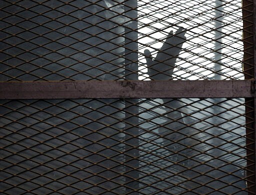 FILE - In this Aug. 22, 2015 file photo, a Muslim Brotherhood member waves his hand from a defendants cage in a courtroom in Torah prison, southern Cairo, Egypt. In some cells in Iran, Egypt, Syria and other countries in the Middle East, prisoners are crammed in by the dozens, with little access to hygiene or medical care. So if one infection gets in, the novel coronavirus could run rampant. (AP Photo/Amr Nabil, File)