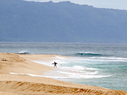 A surfer walks out of the ocean on Oahu's North Shore near Haleiwa, Hawaii, Tuesday, March 31, 2020. The state of Hawaii is reporting its first death of an individual who tested positive for the coronavirus. The state Department of Health says the individual was an older male resident of Oahu. He had been hospitalized recently with multiple medical issues but it's not clear what his cause of death was. He did test positive for the disease and had been exposed to someone who had tested positive. (AP Photo/Caleb Jones)