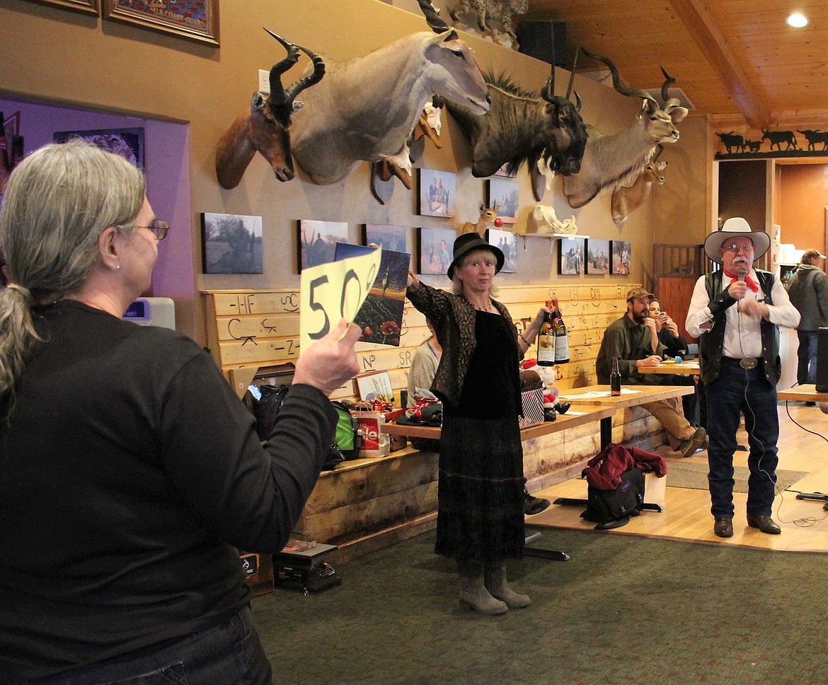 Auction helper Kristi Thistle holds up a painting from a Missoula artist and some wine as long-time auctioneer, D.K. Mitchell, calls for bids. The Helping Hands Auction was held on Dec. 2 at the River Edge in Alberton. (Kathleen Woodford/Mineral Independent)