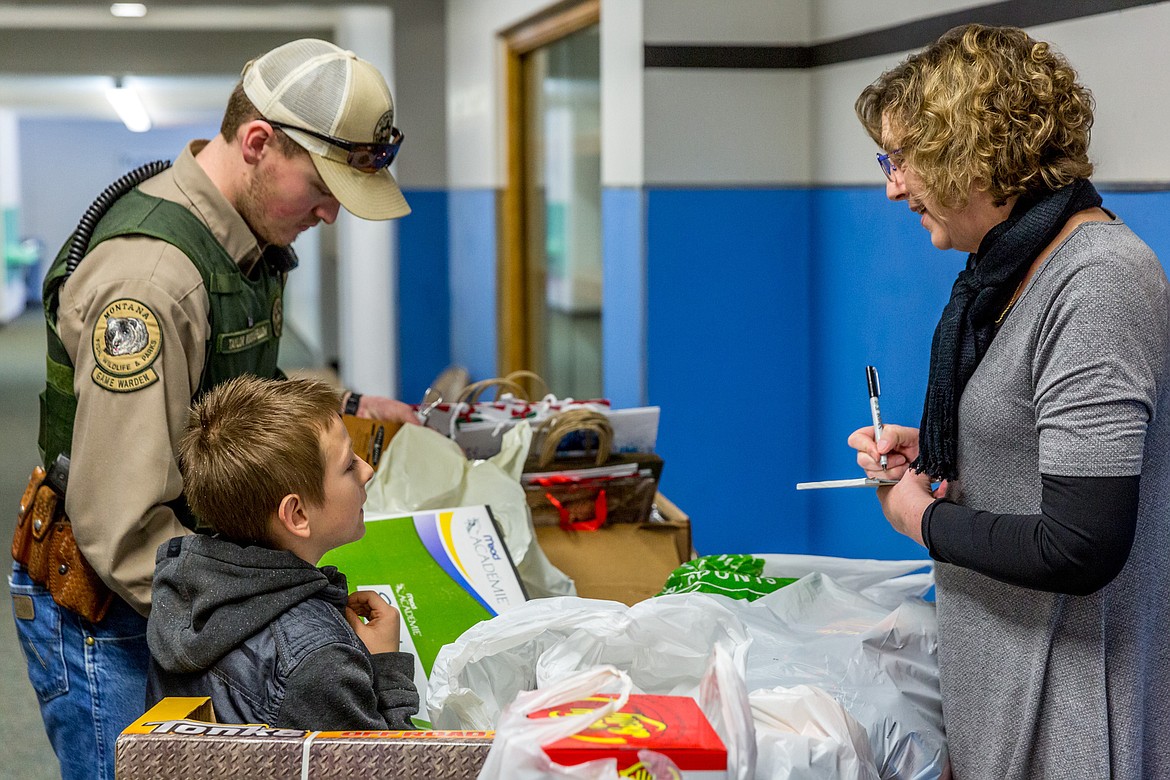 Tyler Simanovicki, 8, of Troy tells Carol Oedewaldt which Christmas gifts go to which family members at a wrapping station in Asa Wood School in Libby Tuesday, Dec. 19, 2017. Simanovicki was participating in Shop With a Cop with Fish, Wildlife and Parks Game Warden Taylor Rockafellow to his left. (John Blodgett/The Western News)