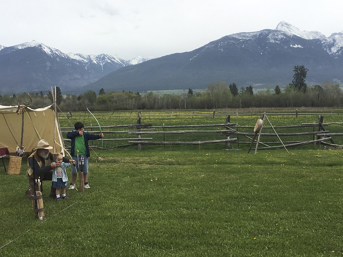 GARY STEELE, left, kneels as he teaches Meadow Mikkelsen, 3, how to shoot a bow and arrow. Behind them is Jamason Mikkelsen, 9. The Mikkelsen family, from Hot Springs, spent the morning at Fort Connah during the annual Rendezvous. (Ashley Fox/Lake County Leader)