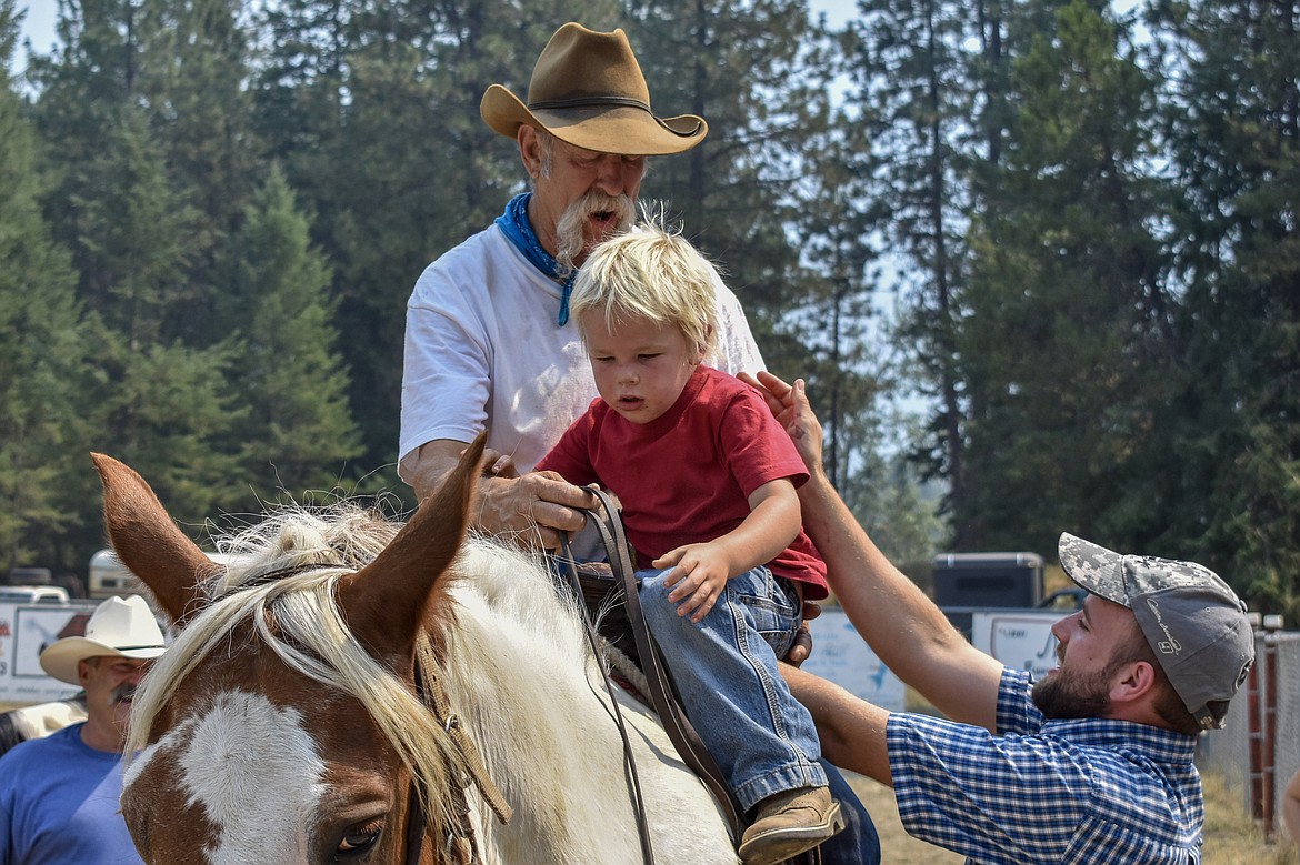 Kevin Lamp helps son Wyatt Lamp onto a horse in front of Gib Stovall for a ride during the Three Lakes Roundup put on by Three Lakes Church near Troy on Saturday. (Ben Kibbey/The Western News)