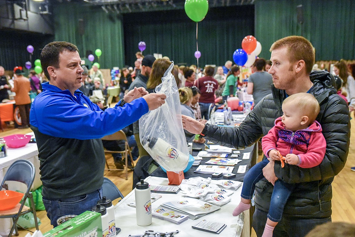 Libby Public Schools Superintendent Craig Barringer -- working the Best Beginnings Coalition table -- hands some free items to Michael Wells -- holding Claire Wells -- at the Cabinet Peaks Medical Center Annual Health Fair at the Libby Memorial Events Center on Saturday. (Ben Kibbey/The Western News)