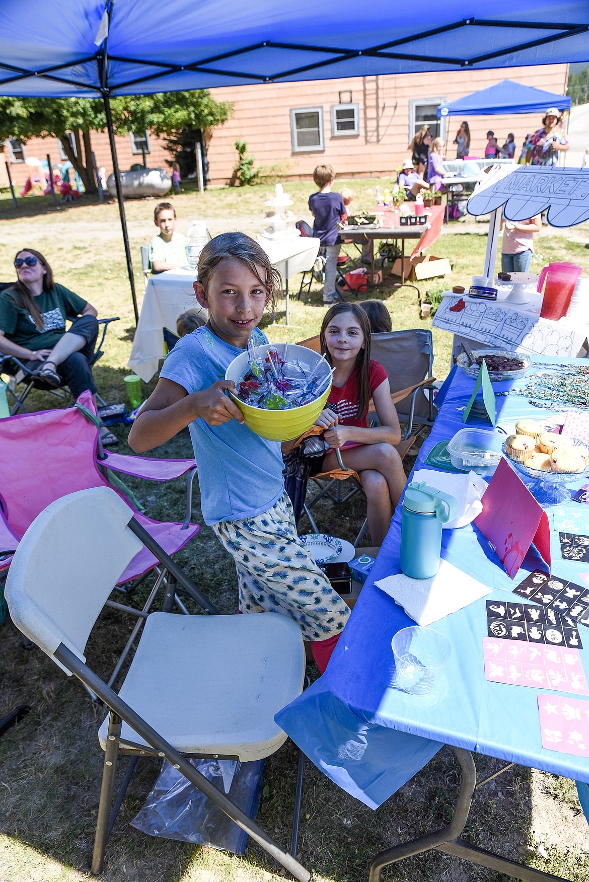 Sammy Evans holds up a bowl of homemade candy she and friend Jenni Brumbaugh (background) were selling at the Libby Kids' Market next to Hotel Libby, Saturday. Evans also had huckleberry muffins, face painting stencils and bandanas she sewed.  (Ben Kibbey/The Western News)