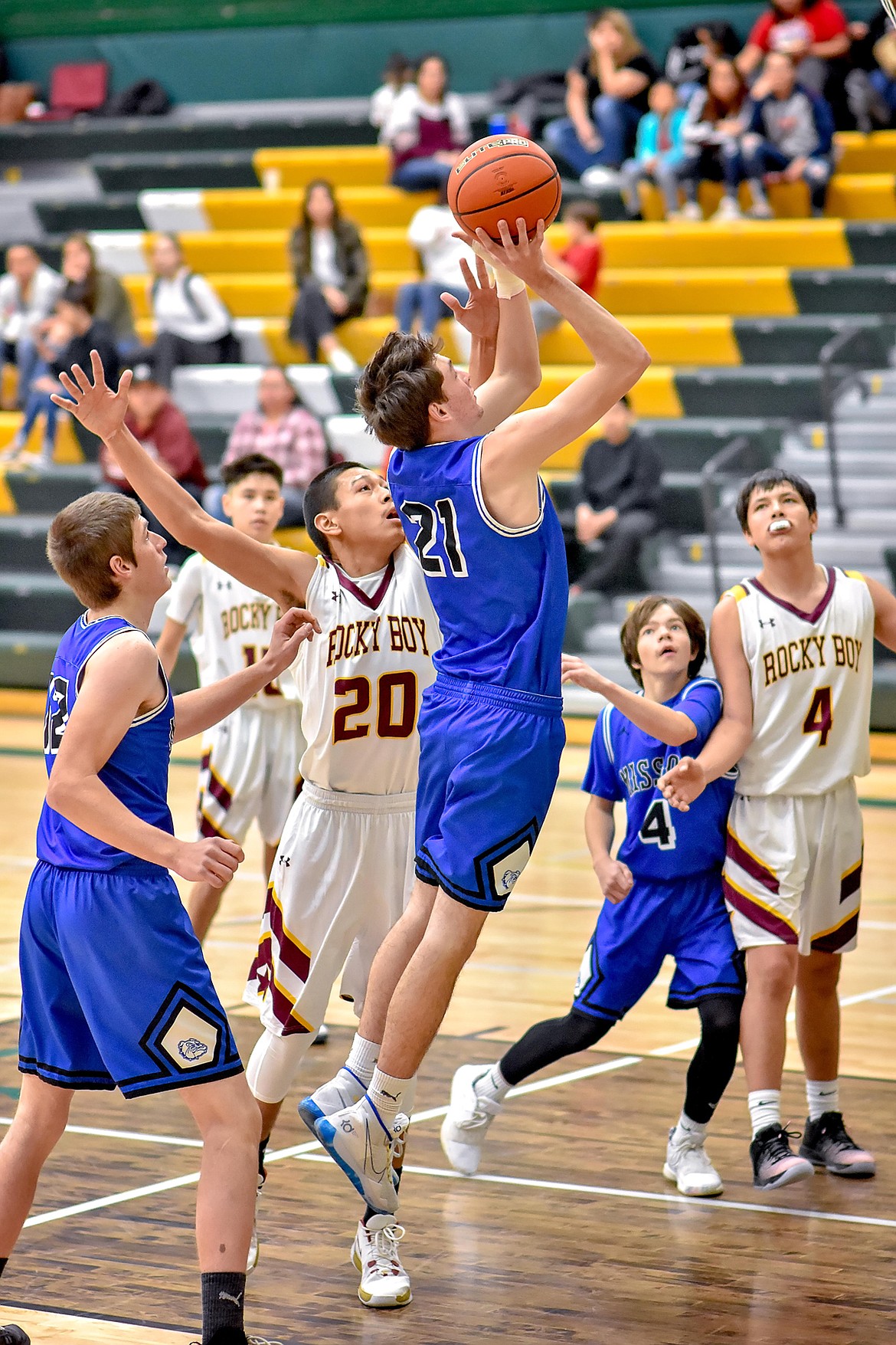 Mission Bulldog Ayden Rosenbaum (21) launches a shot against the Rocky Boy Stars at the Native American Classic in Great Falls, while Ross McPherson (42) positions for a rebound and (4) Zoran LaFrombois boxes out his opponent. (Photo courtesy of Christa Umphrey, Forward Photography)