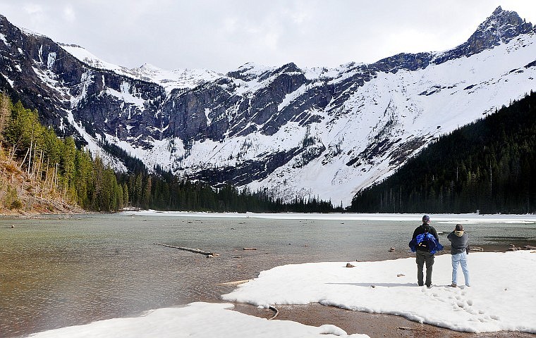 Two people stand in snow on the beach while admiring the view at Avalanche Lake in Glacier National Park on April 25. Portions of roads and trails in the park are open despite recent spring snowfall.